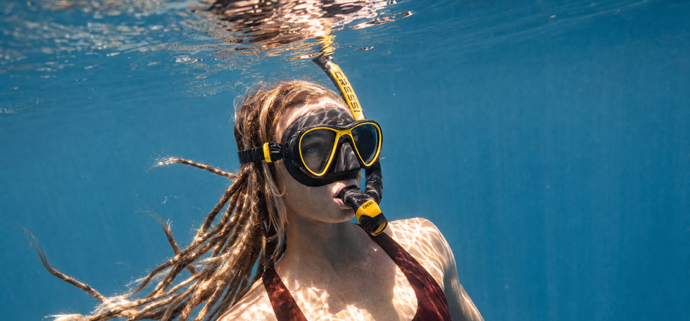 A woman underwater wearing a black and yellow snorkel mask with a snorkel, a dark red swimsuit, and her hair floating in the blue water.